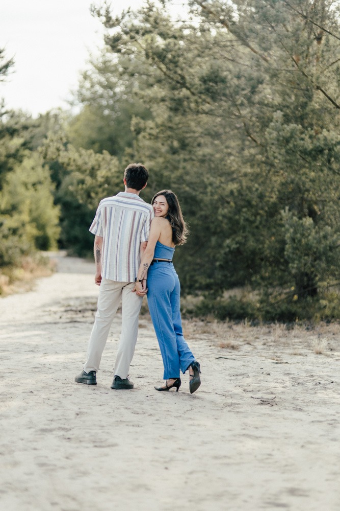 Ein Paar hält sich an der Hand und schaut sich an, während sie auf einem sandigen Weg durch einen Wald gehen.